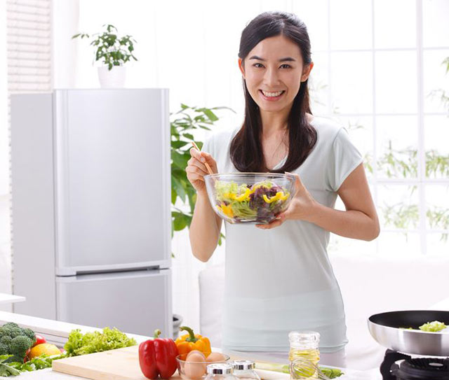 Photo of a woman standing behind kitchen counter holding a glass bowl of salad